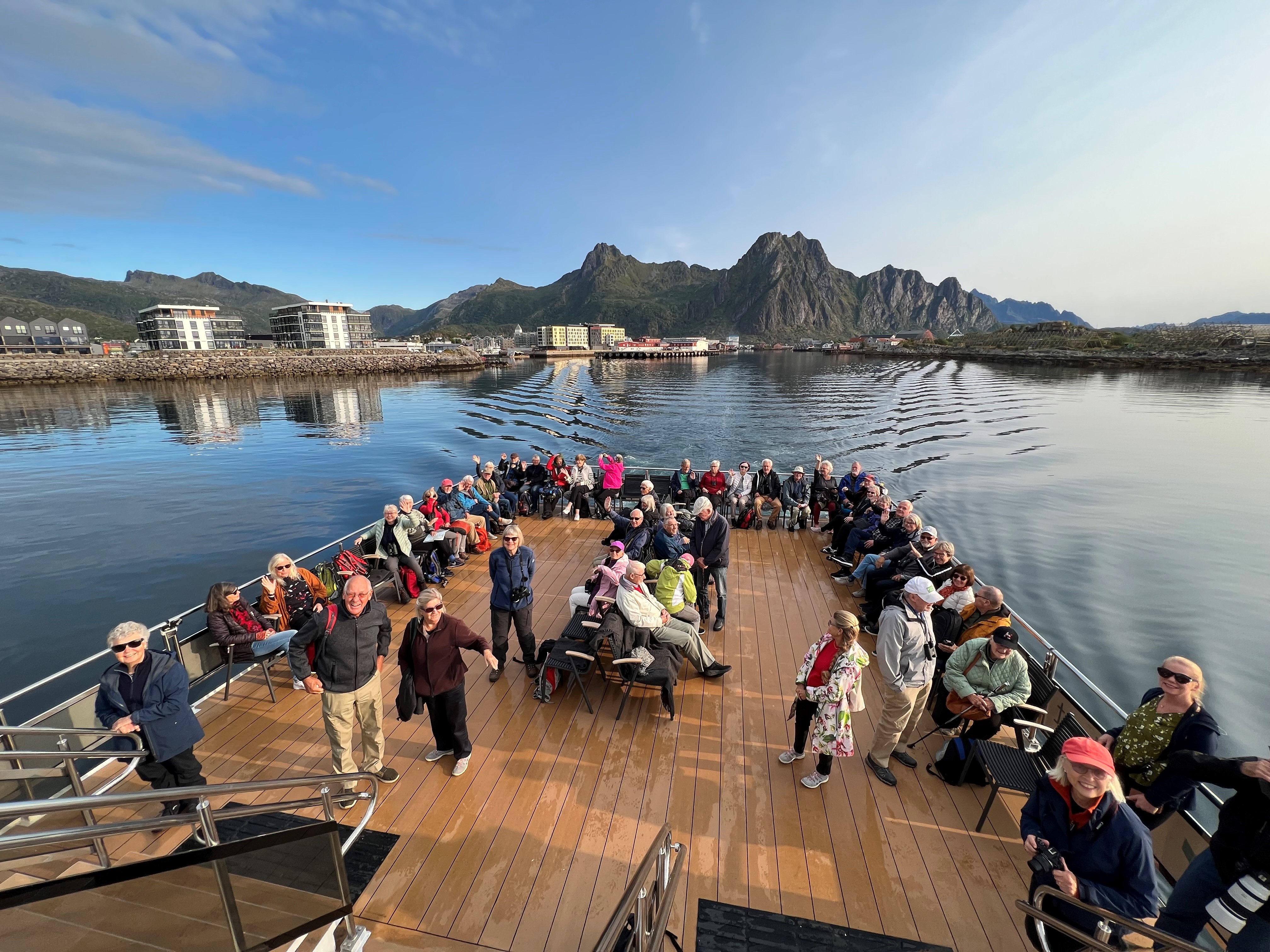 På bilden ser vi en båt med stort utomhusdäck med passagerare och berg och hav i bakgrunden